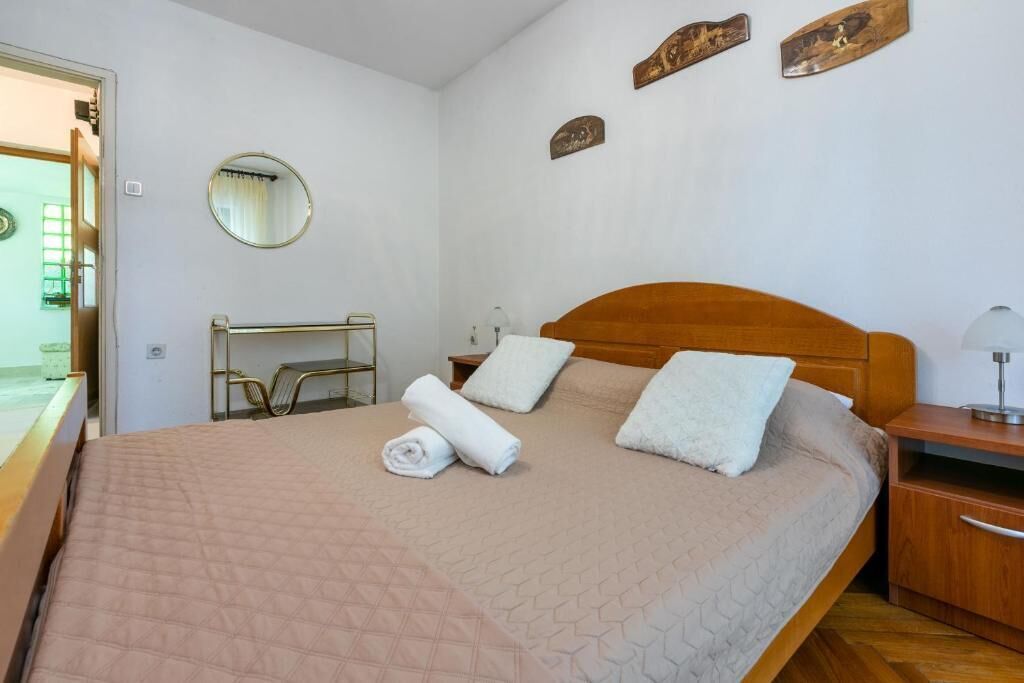 Una cama bien hecha con cabecera de madera, una mesita de noche con lámpara y un pequeño estante de madera.