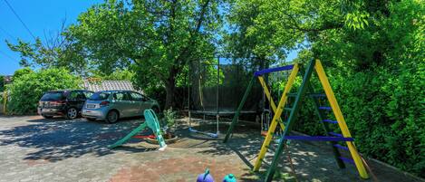 A playground with a swing set, a slide, and a purple spinning ride. There are two cars parked nearby.