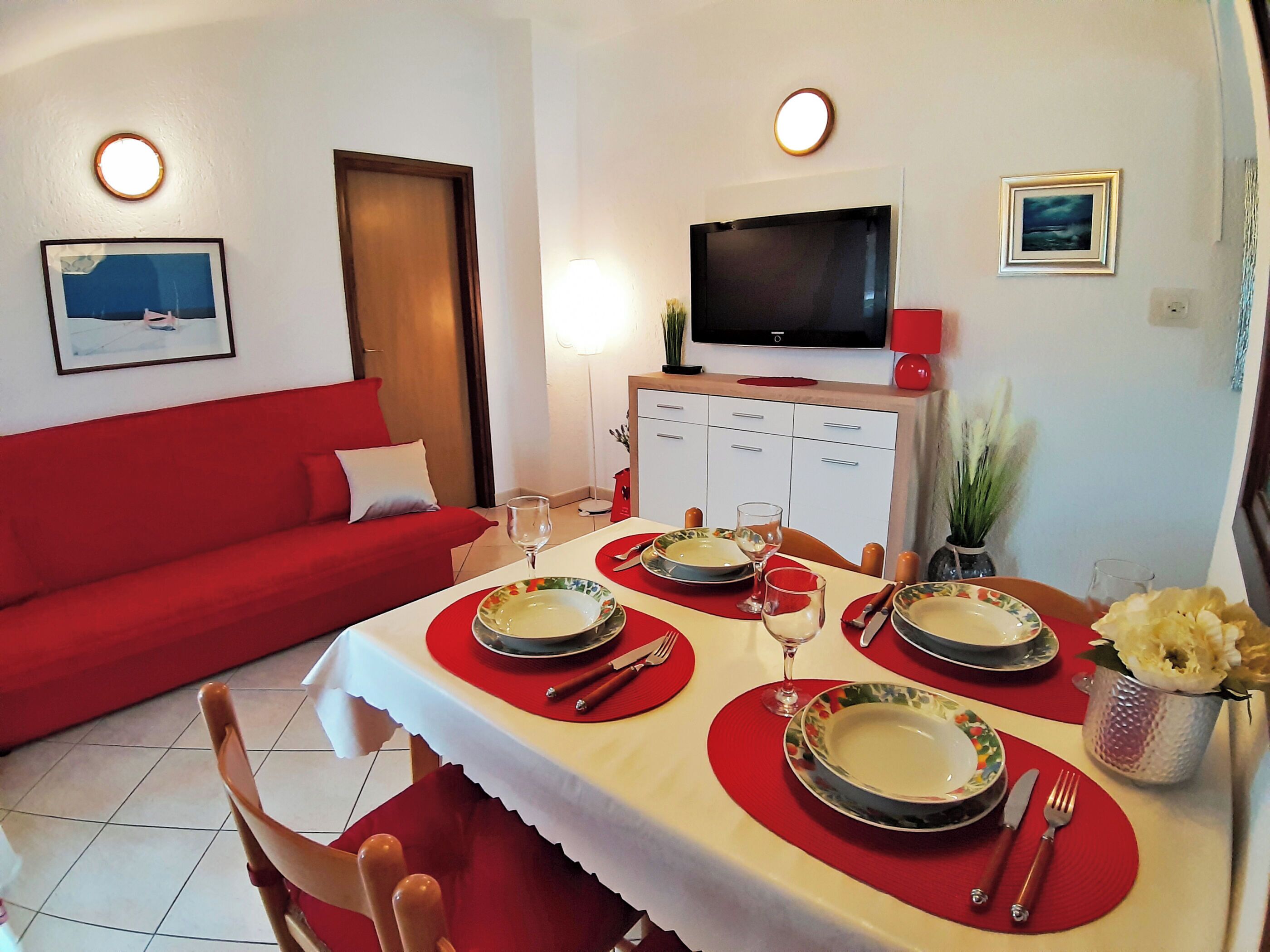 A dining area with a table set for four, a red sofa, a TV, and a small plant on the wall.