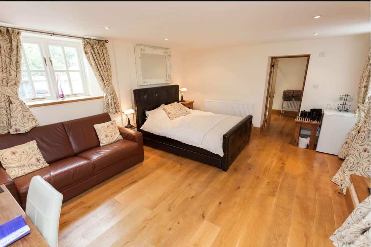 A bedroom with a bed, a brown leather sofa, a wooden floor, a window with curtains, a mirror, and a small kitchenette area.
