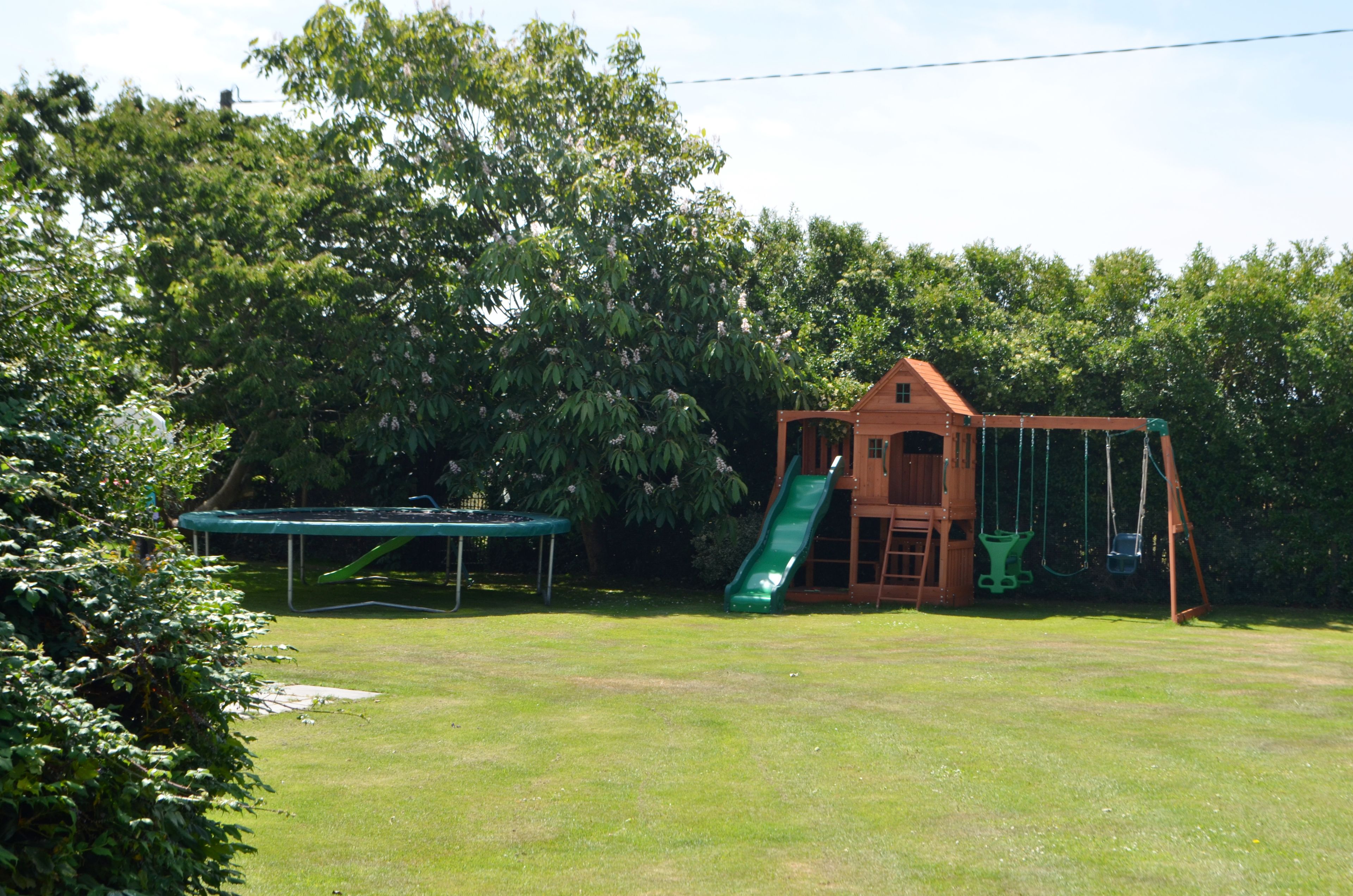 A wooden playhouse with a slide and swings, surrounded by a grassy yard and trees.