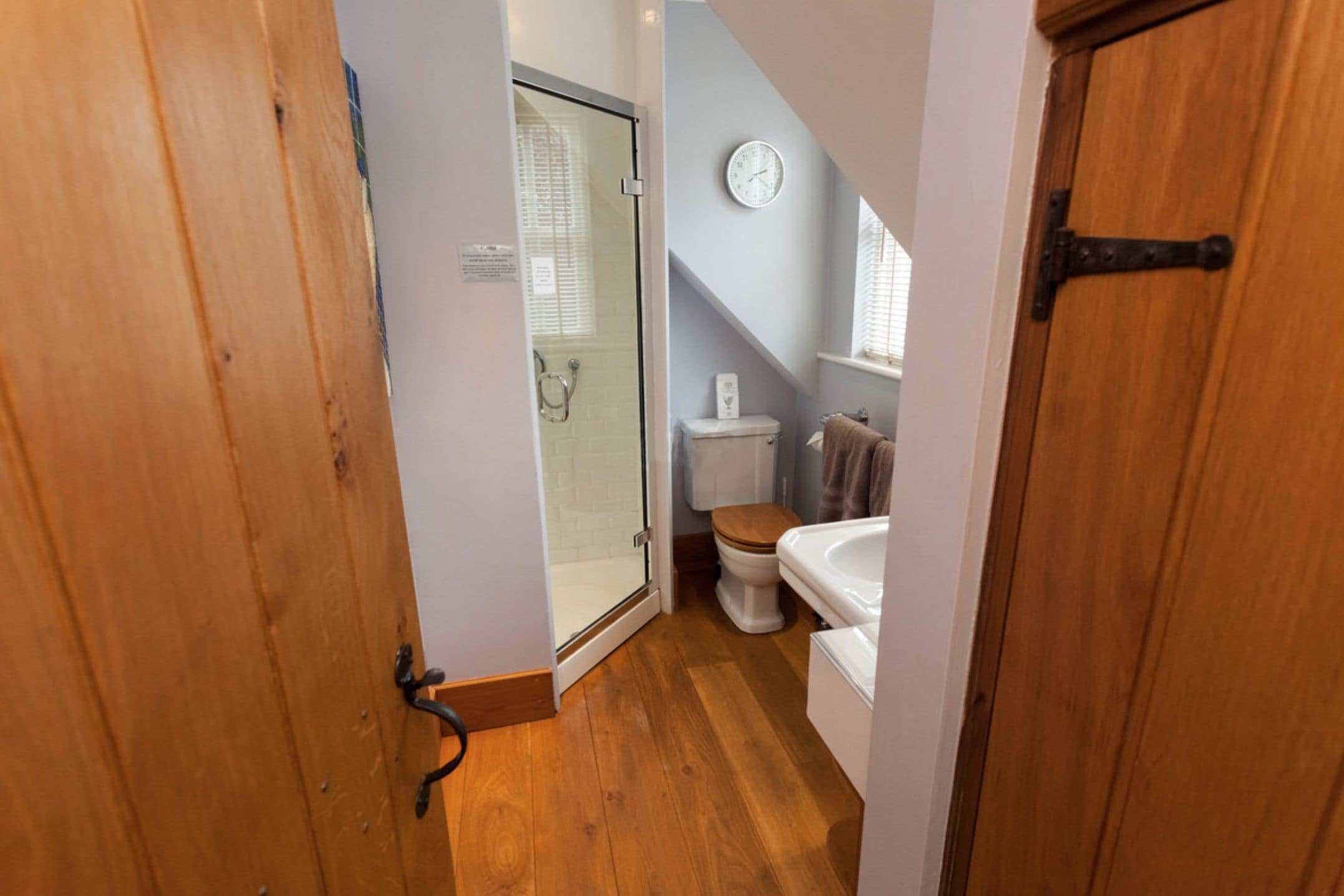 A narrow bathroom with a wooden door, a glass shower enclosure, a toilet, and a sink.