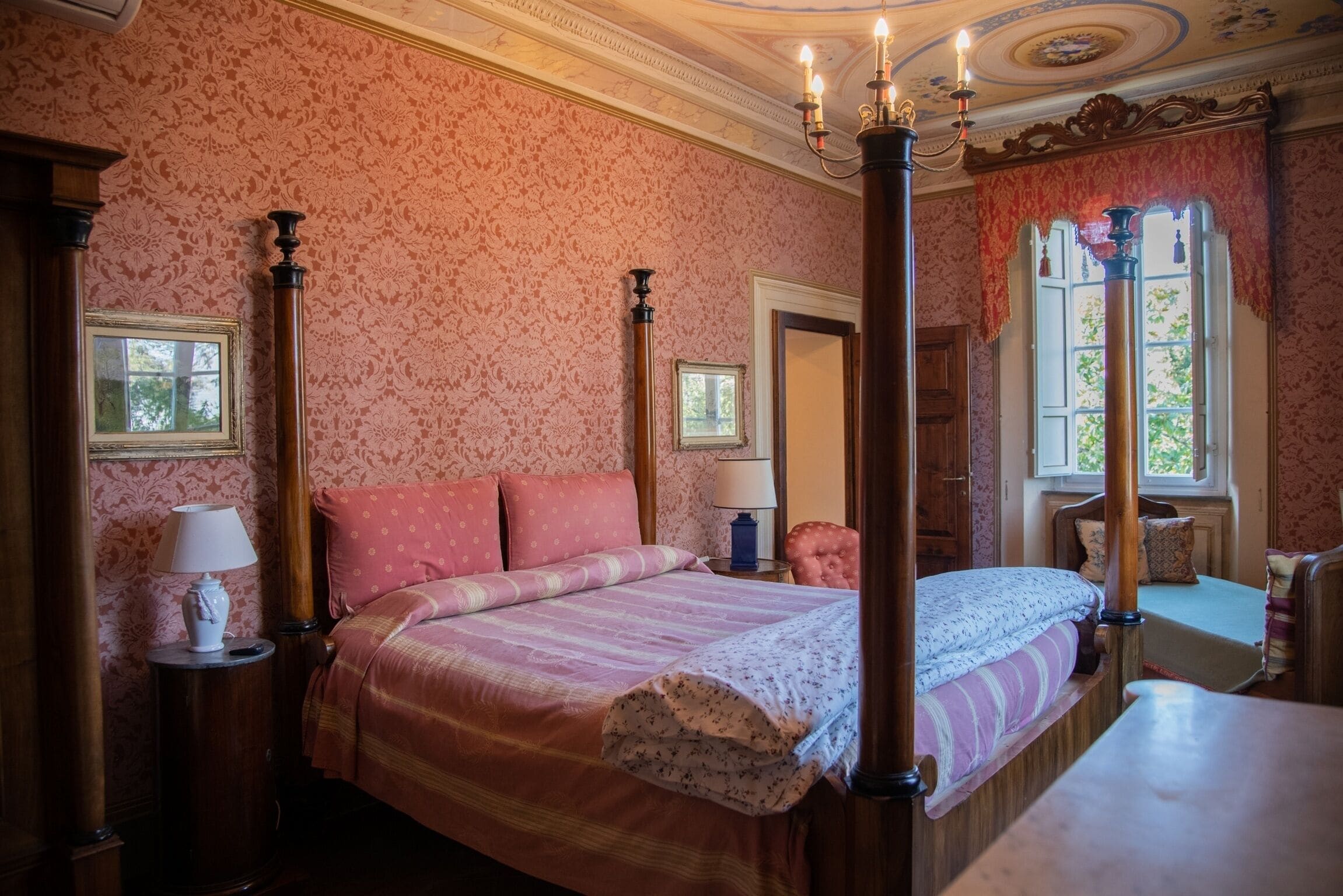 A four-poster bed with a floral bedspread, a nightstand with a lamp, and a window with a view of greenery.