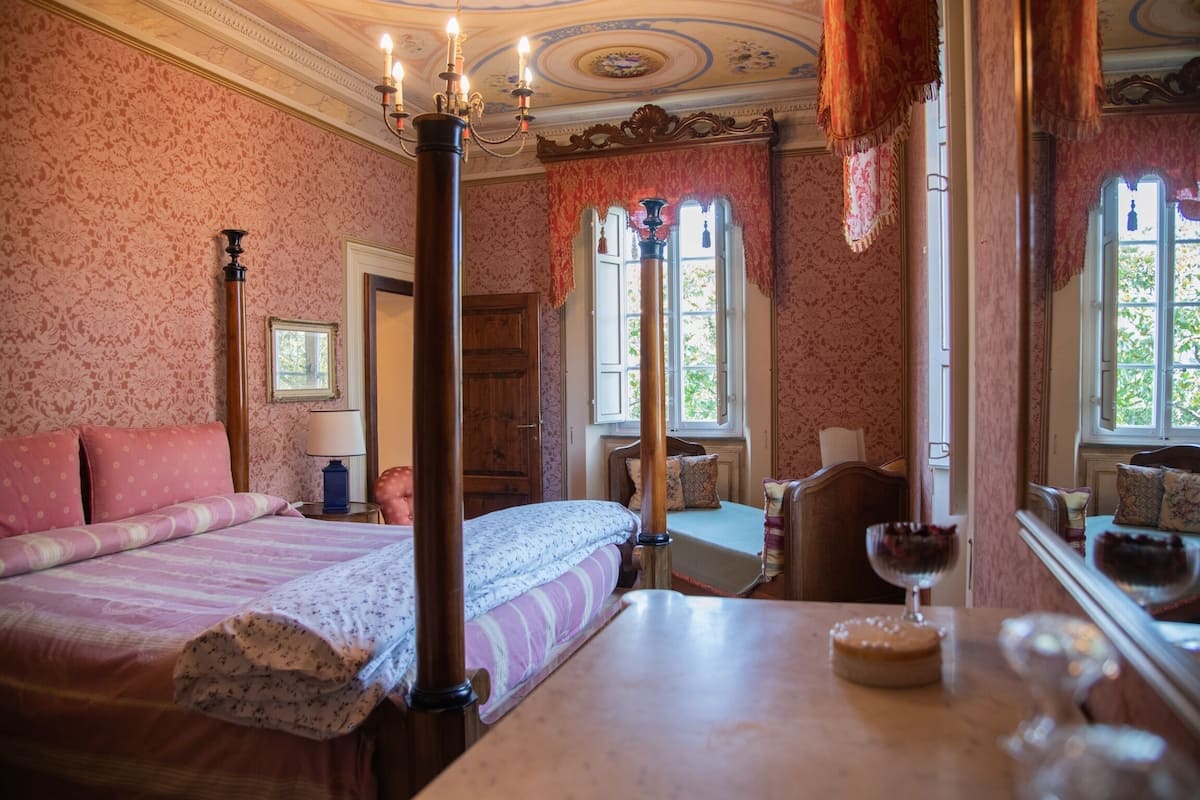 A bedroom with a four-poster bed, a wooden table with a glass, and a mirror reflecting the room.