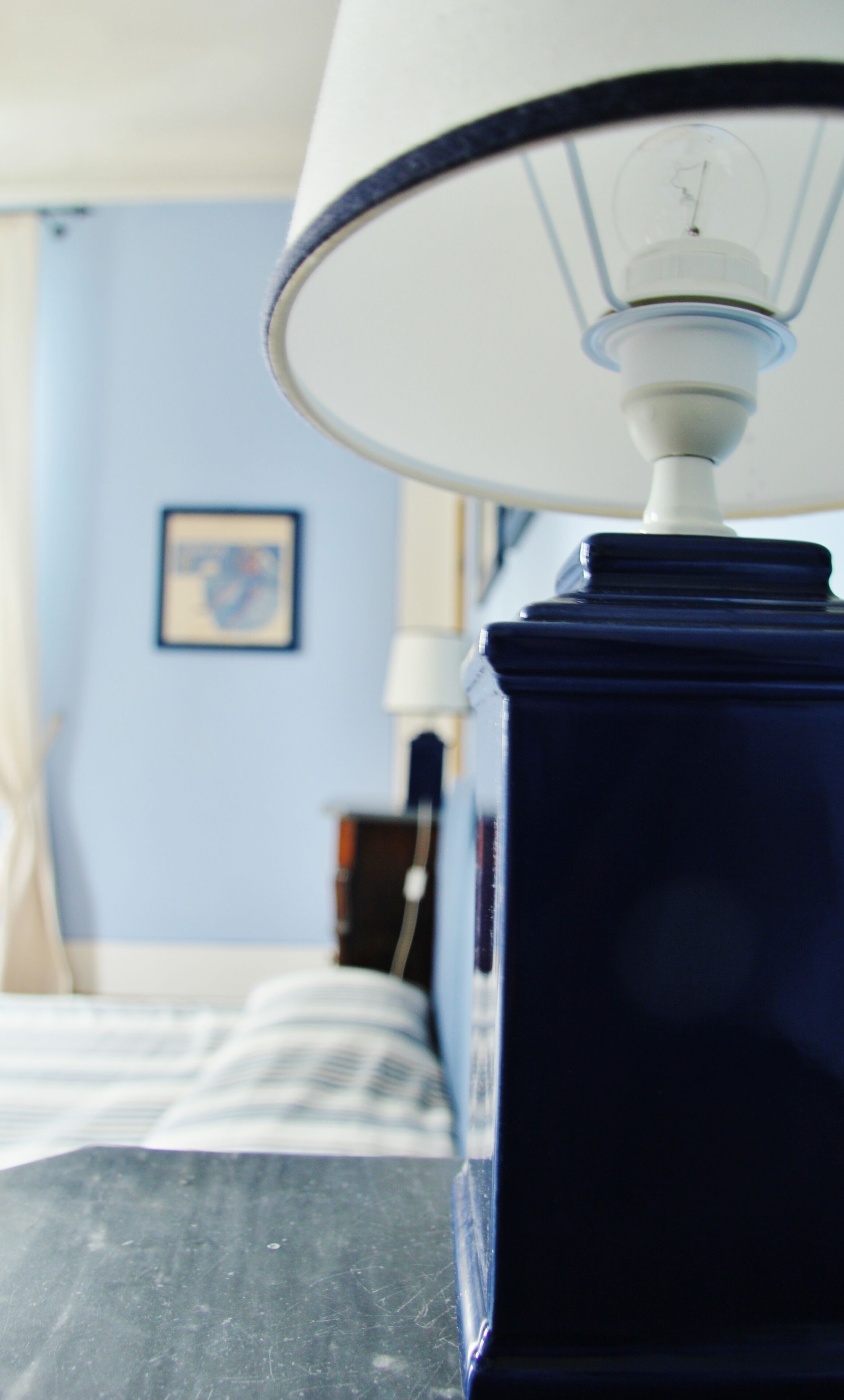 Eine Nahaufnahme einer blauen Tischlampe mit weißem Lampenschirm, platziert in einem Zimmer mit einem Bett und einem gerahmten Bild an der Wand.