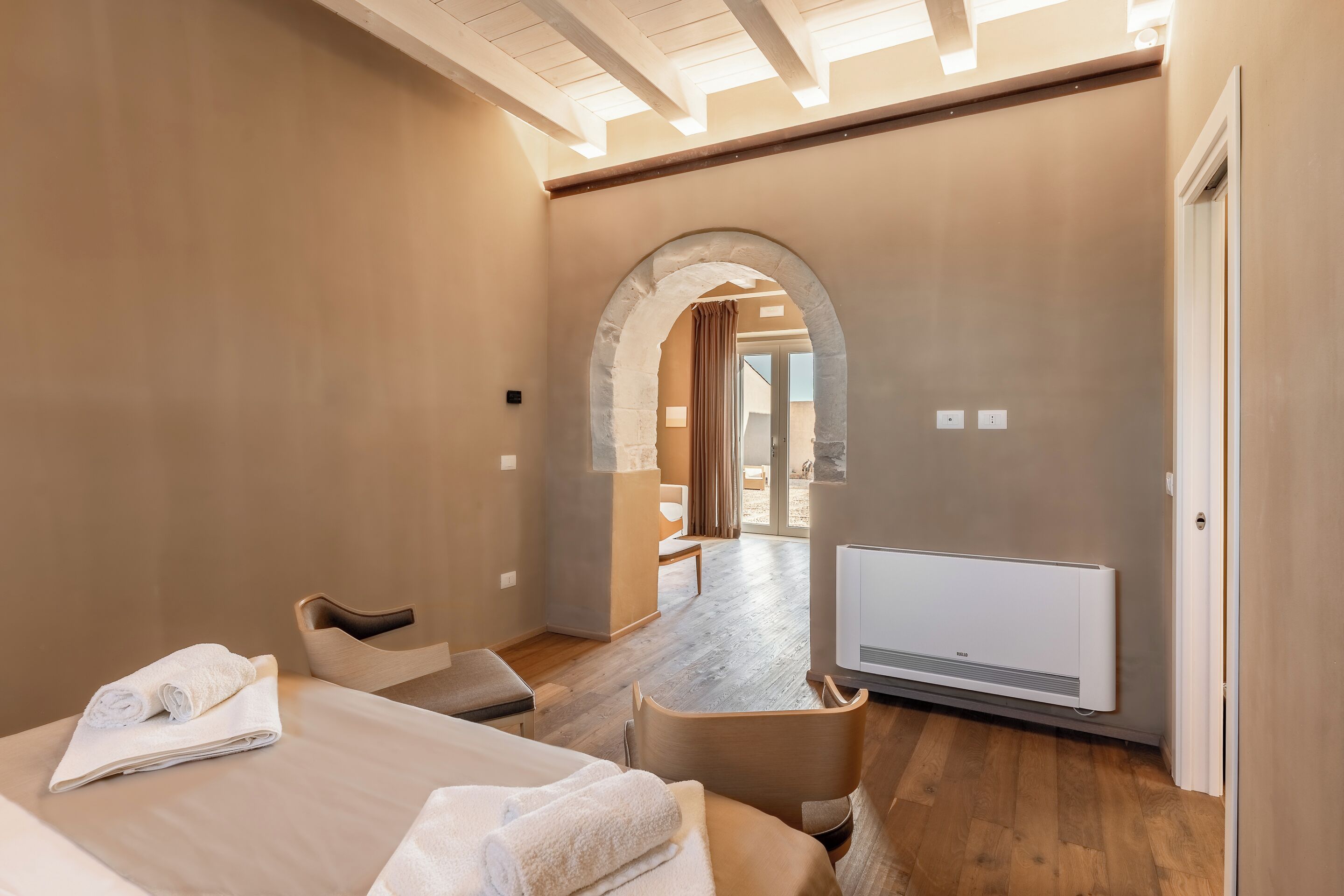Una habitación de hotel moderna con piso de madera, una cama con ropa blanca, una zona de estar y vistas a un balcón a través de un arco.