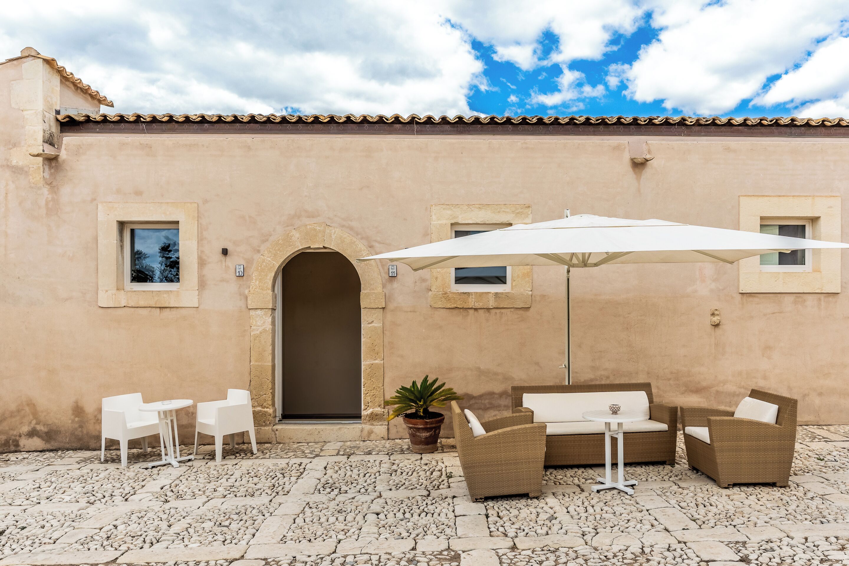 An outdoor patio with a beige wall, a tiled floor, white chairs, a sofa, and a large umbrella.