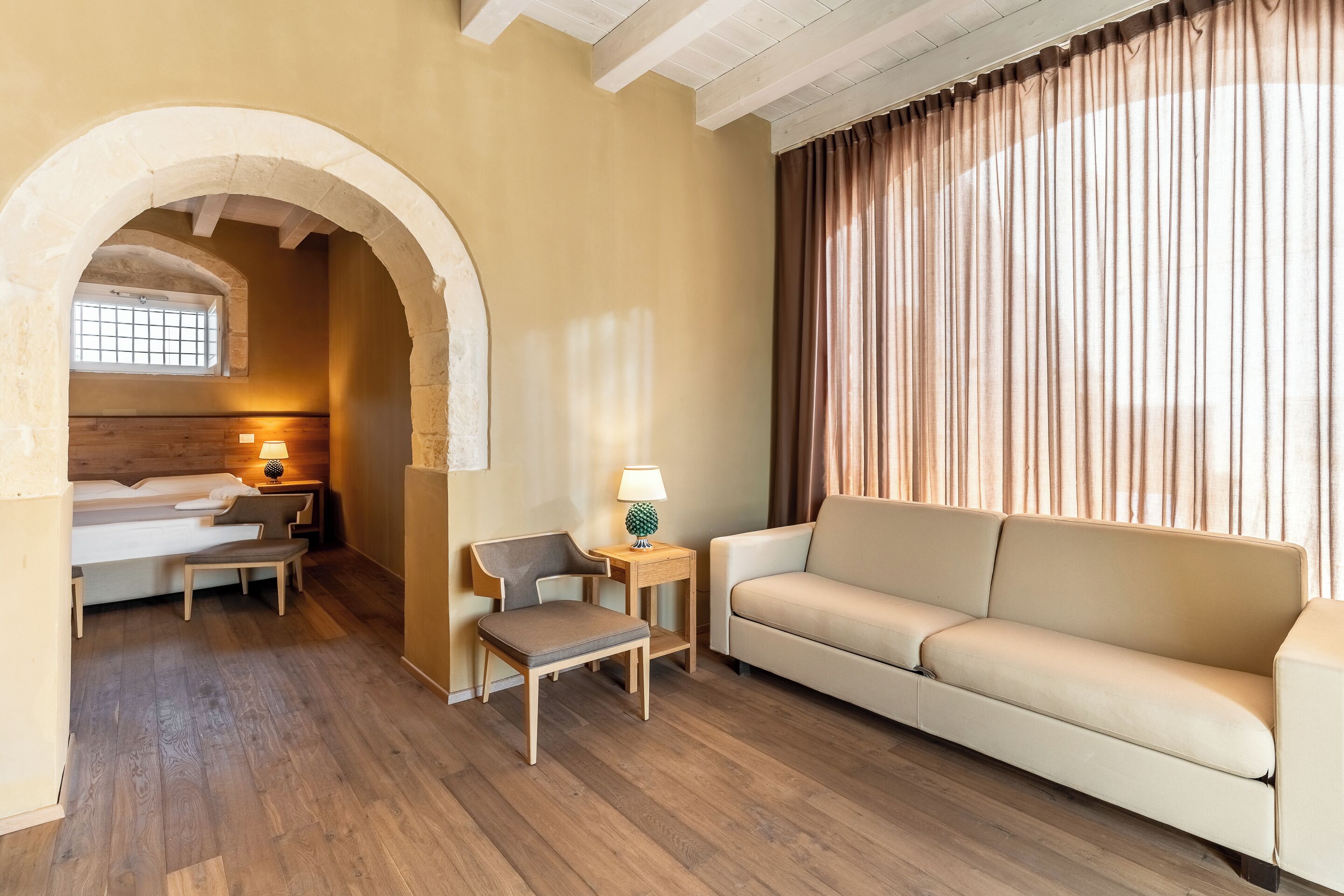 Hotelkamer met houten vloer, een bank, een kleine tafel met lamp, een bed met hoofdeinde en een raam met gordijnen.