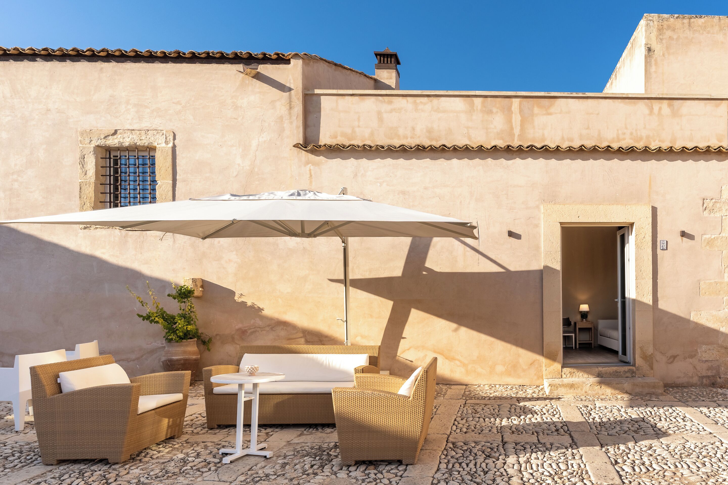 An outdoor seating area with wicker furniture and a white umbrella, set against a building with a tiled roof and stone walls.