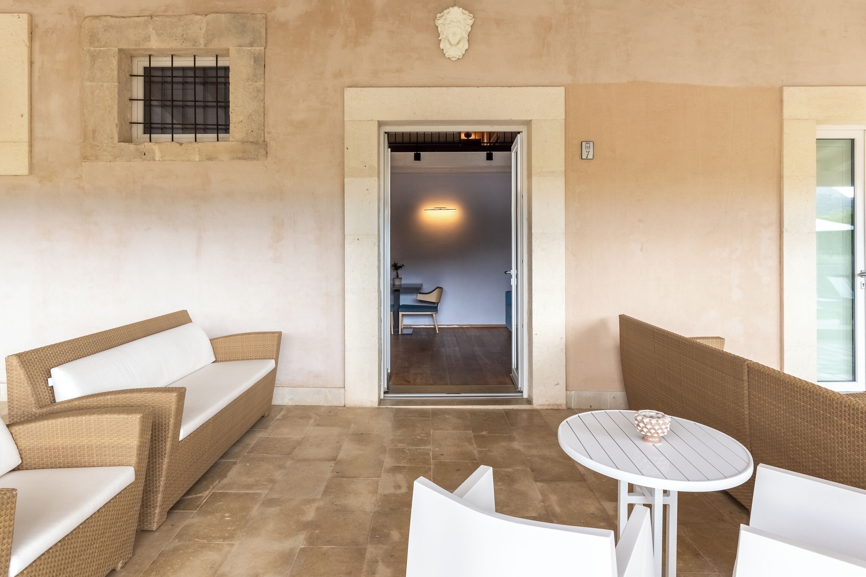 Una stanza con una parete beige, una finestra con cornice in pietra, un divano bianco, una sedia in vimini e un tavolo rotondo con piano bianco.