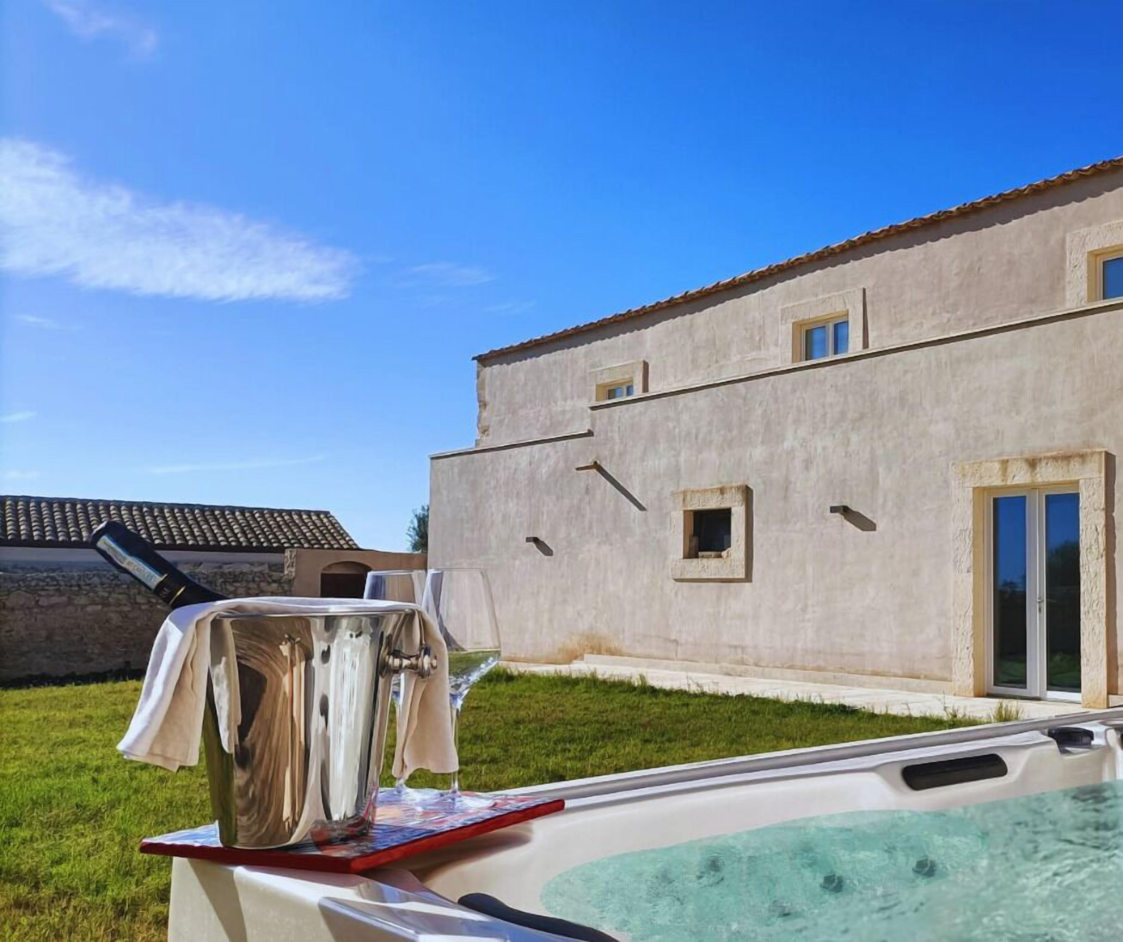 Ein Whirlpool mit Glasabdeckung, eine Flasche Wein, ein Tisch mit Tischdecke und im Hintergrund ein Gebäude mit Ziegeldach.