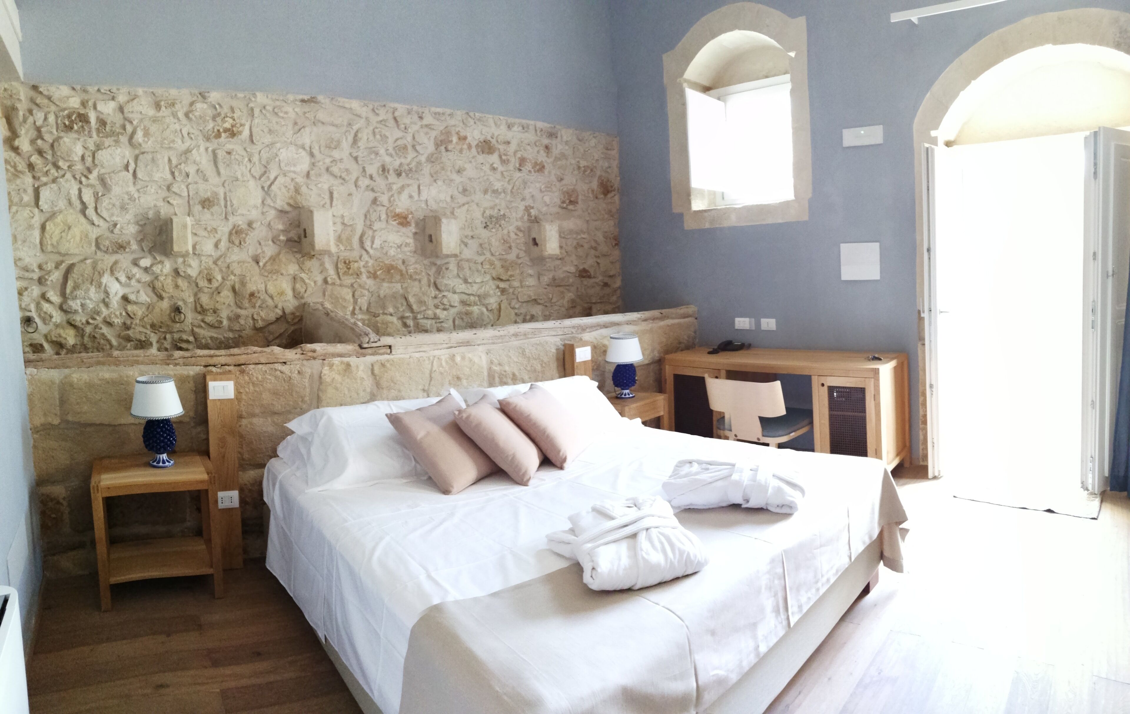 Un dormitorio con pared de piedra, una cama con ropa de cama blanca, dos mesitas de noche con lámparas, un escritorio de madera y un espejo.