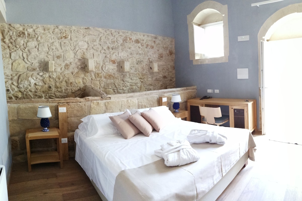 Un dormitorio con pared de piedra, una cama con ropa de cama blanca, dos mesitas de noche con lámparas, un escritorio de madera y un espejo.