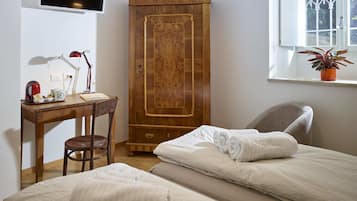 Une chambre avec deux lits, une armoire en bois, un bureau avec une chaise, un téléviseur fixé au mur et une fenêtre avec une plante.