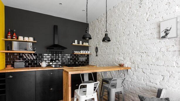 A modern kitchen with a black and white color scheme, featuring a wooden countertop, black cabinets, and a brick backsplash. There are two bar stools and a small dining table. - Urban Rustic Flat in Historical  Building (Śródmieście)