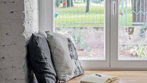 A room with a white brick wall, a window with a view of a green park, a wooden bench with pillows and a book, and a small potted plant. - Urban Rustic Flat in Historical  Building (Śródmieście)