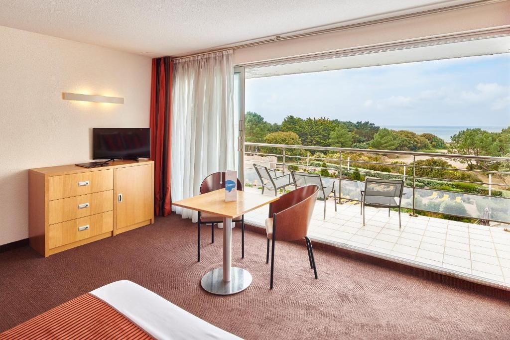 A hotel room with a bed, a TV, a desk, a chair, and a balcony with a view of the sea and greenery.