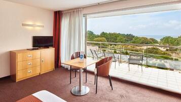 A hotel room with a bed, a TV, a desk, a chair, and a balcony with a view of the sea and greenery.