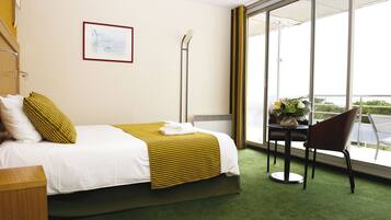 A hotel room with a bed, a chair, a table with a vase, and a window with a view.