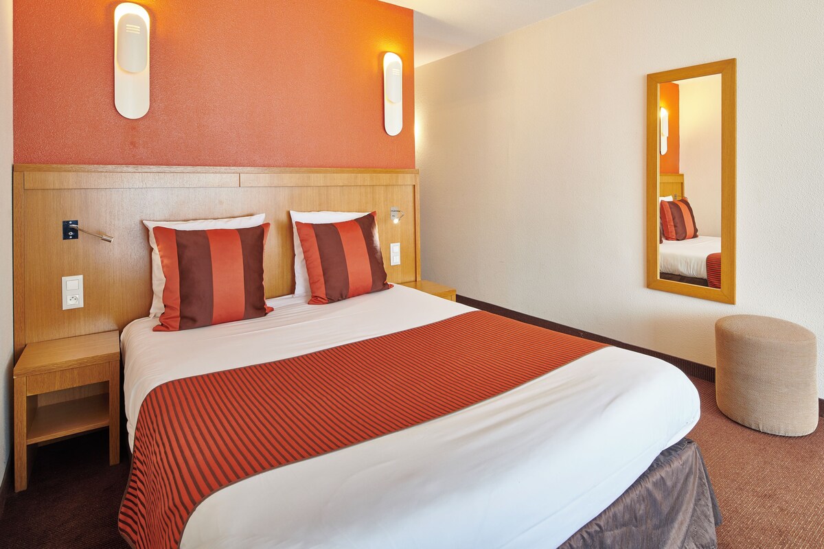 Quarto de hotel com cama, mesas de cabeceira, espelho e luminária na parede.