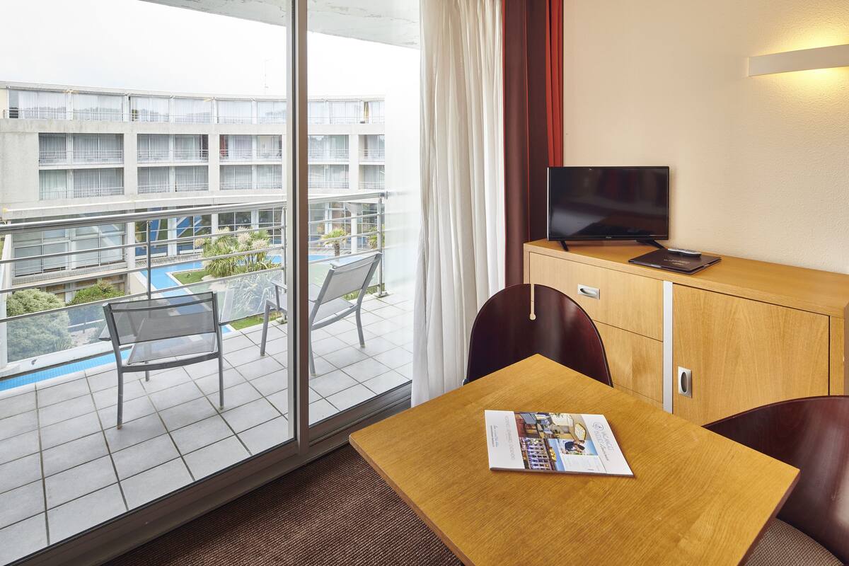 Et hotelværelse med balkon, fjernsyn, et træbord og et magasin.