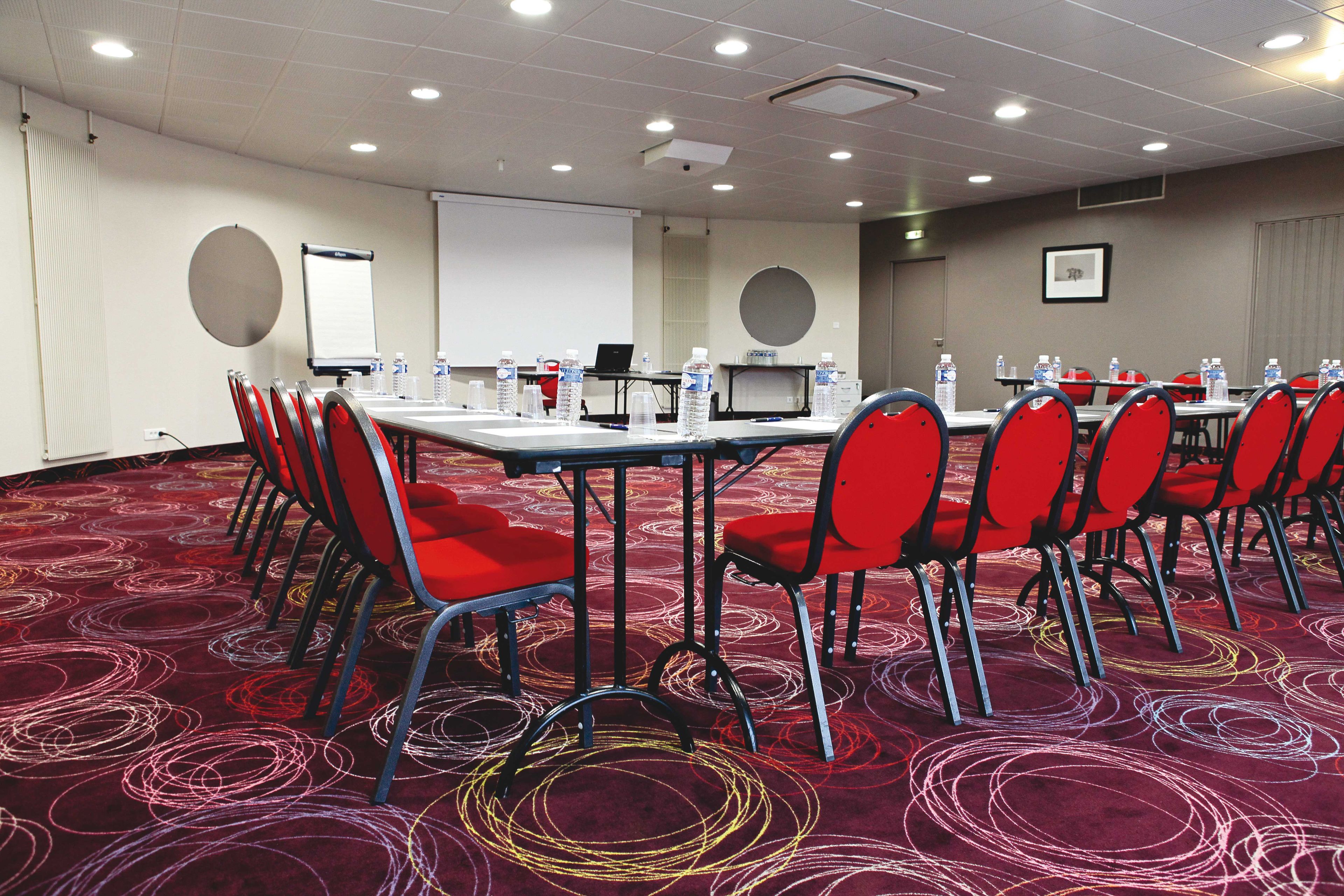 A conference room with a carpeted floor, red chairs, a table with water bottles, a projector screen, and a laptop.