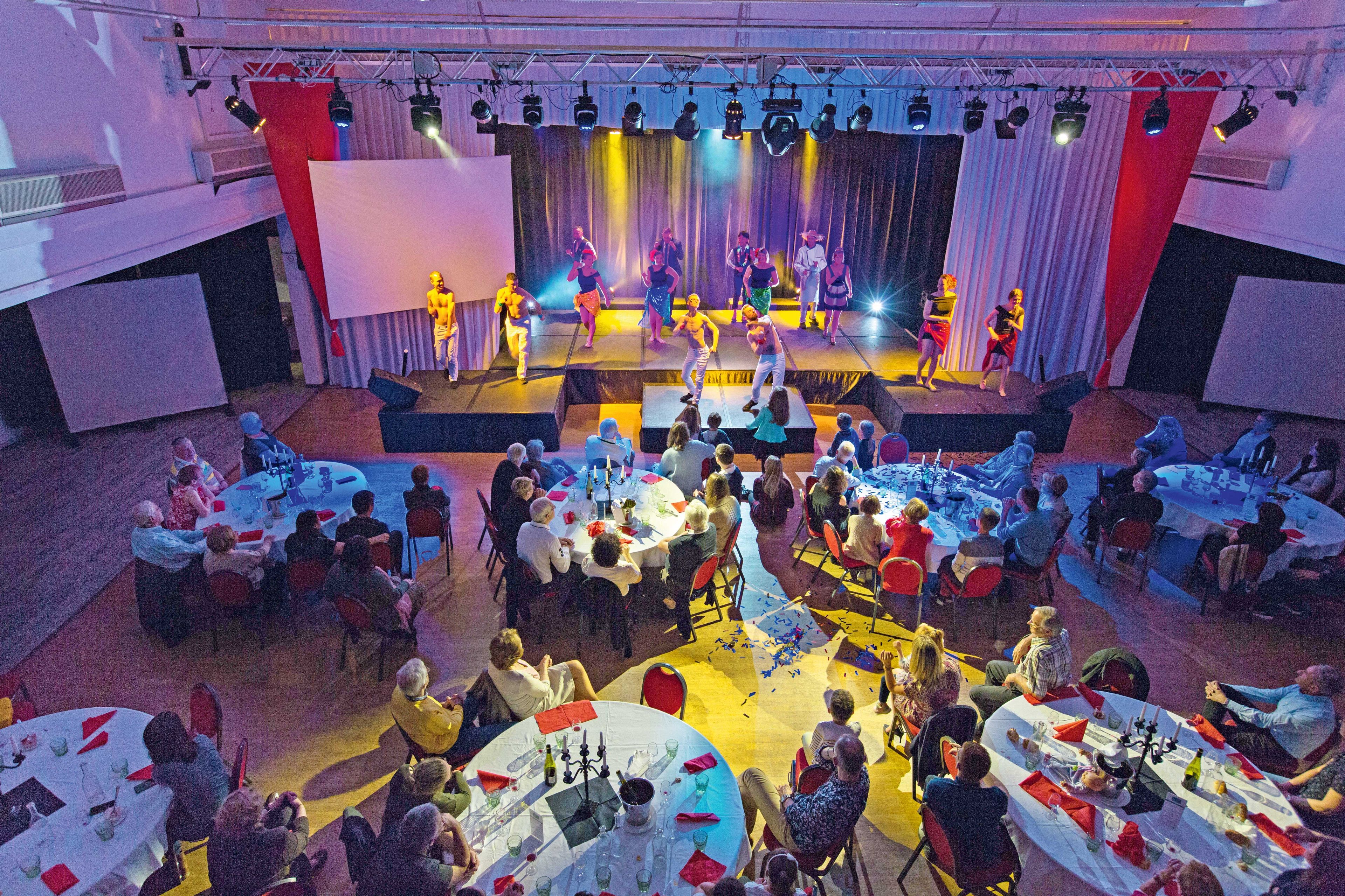 A banquet hall with round tables set for dining, a stage with performers, and a large audience seated.