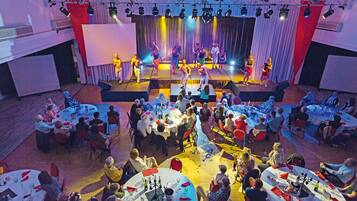 A banquet hall with round tables set for dining, a stage with performers, and a large audience seated.