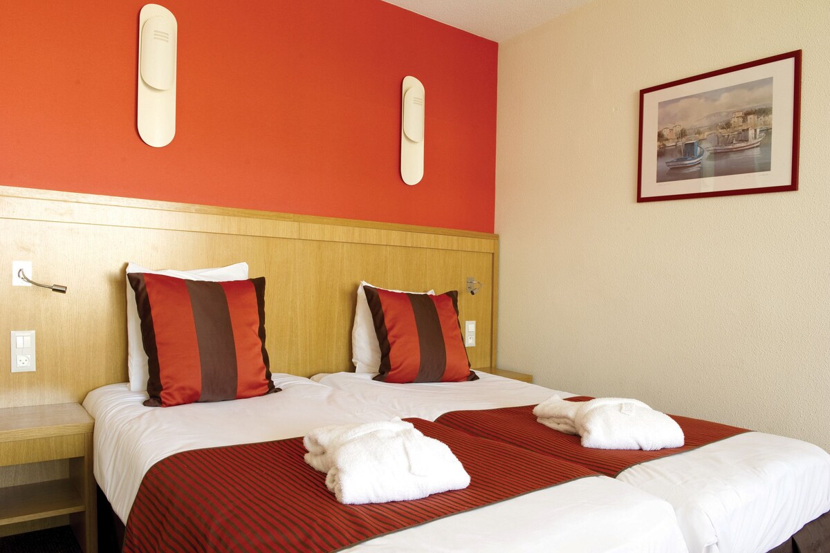 Quarto de hotel com cabeceira de madeira, duas camas com roupa de cama vermelha e branca, um tapete listrado em vermelho e branco e uma moldura com uma imagem na parede.