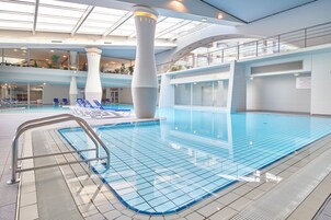 En modern inomhuspool med kristallklart blått vatten, ett tak som släpper in mycket ljus och ett område vid poolkanten med solstolar.