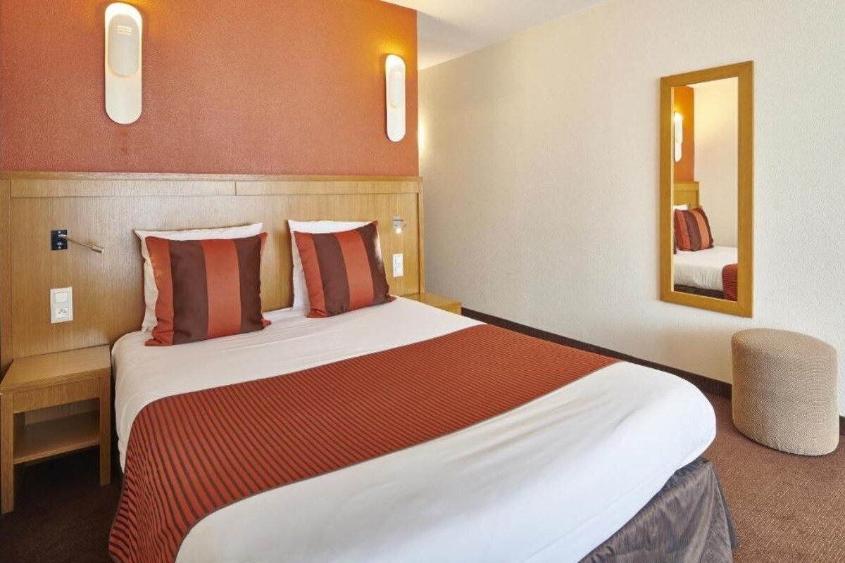 Quarto de hotel com uma cama grande, duas mesas de cabeceira, um espelho e um pequeno banquinho.