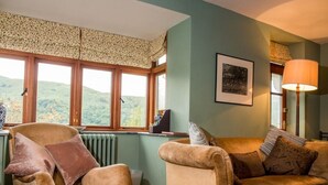 Un salon chaleureux avec vue sur des collines verdoyantes, un radiateur, une lampe et un tableau encadré au mur.
