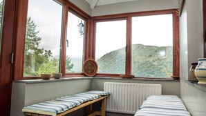 Une pièce avec un banc et un lit, de grandes fenêtres donnant sur des collines, et un radiateur.