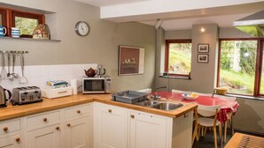 Une cuisine avec des meubles blancs, un four à micro-ondes, une bouilloire et un grille-pain. Une fenêtre offre une vue sur la verdure environnante.