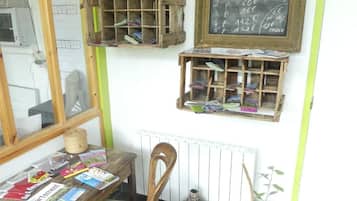 Una habitación con una mesa de madera que muestra libros y revistas, una ventana con persianas, un radiador y un menú de pizarrones con precios.