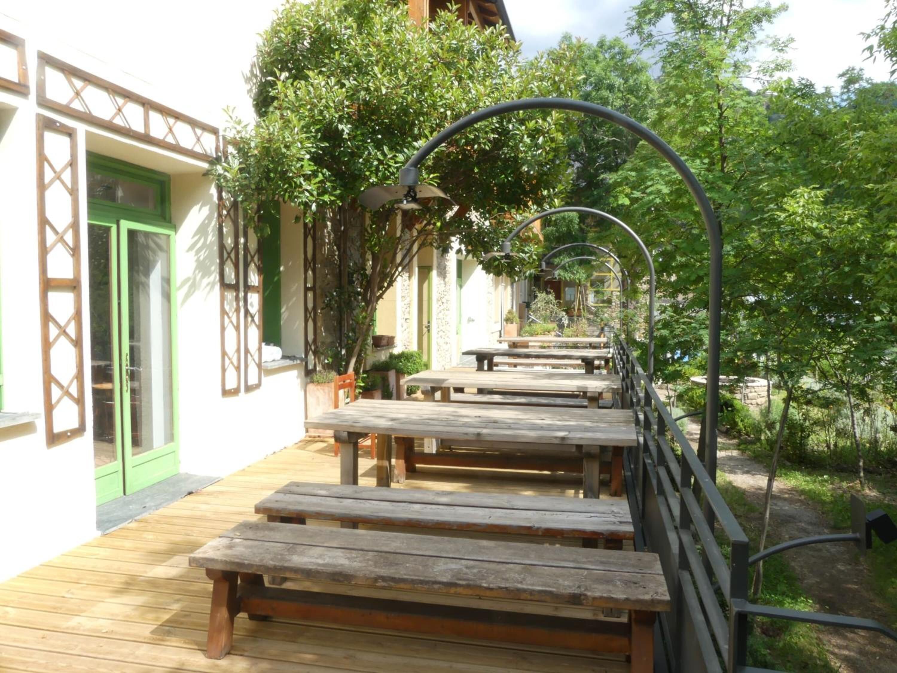 An outdoor patio with wooden benches and tables, surrounded by greenery and a building with a green door and white walls.