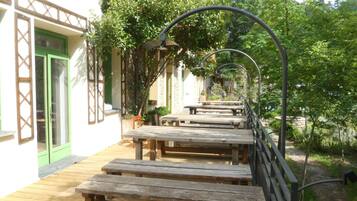 An outdoor patio with wooden benches and tables, surrounded by greenery and a building with a green door and white walls.