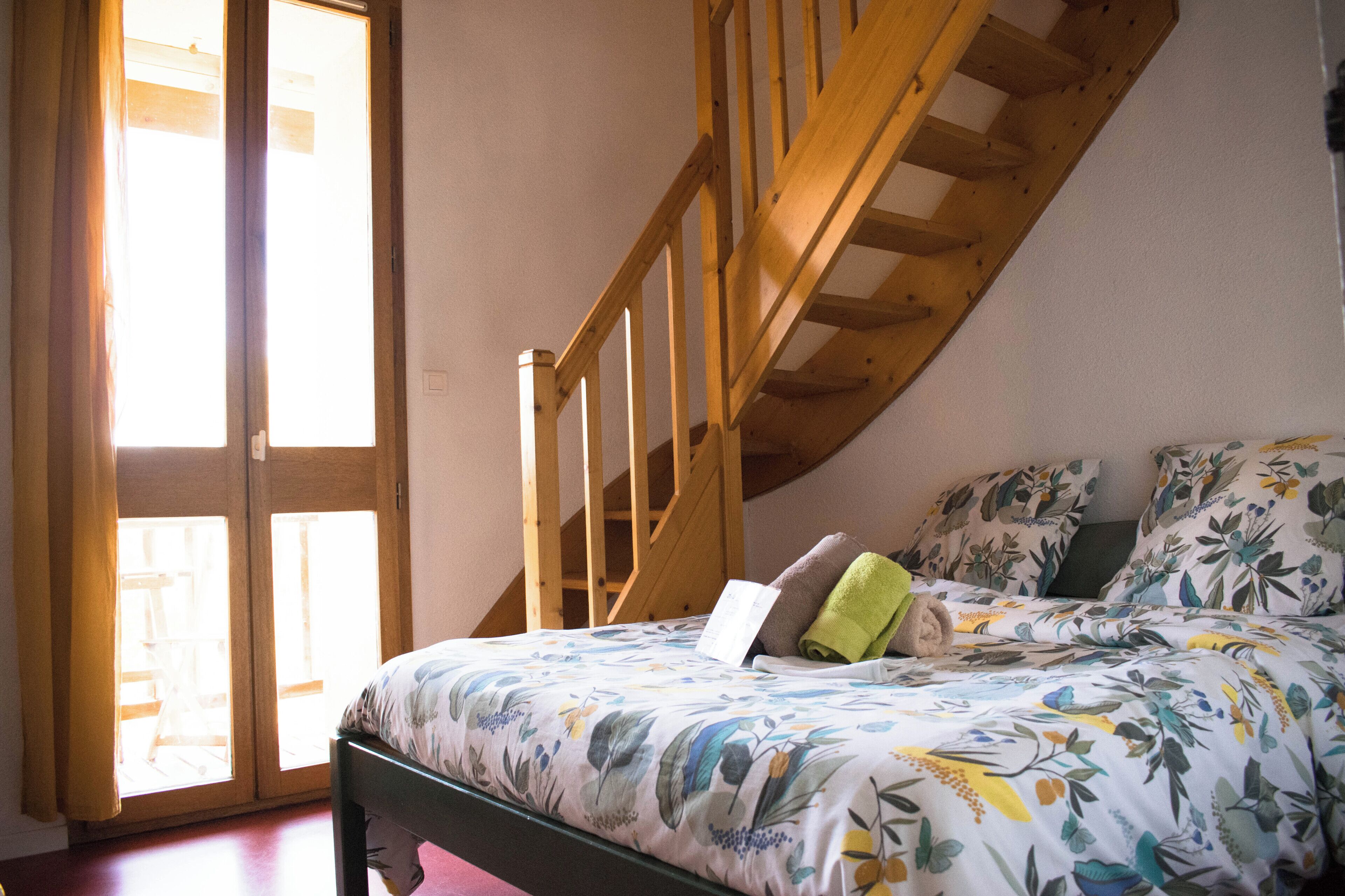 Ein Schlafzimmer mit schrägem Dach, einer Holztreppe, einem Bett mit floralem Bettzeug und einem Fenster mit Vorhängen.