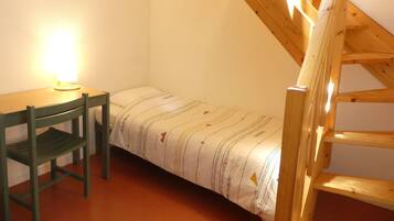 A small, single-bed room with a wooden ladder, a desk with a chair, and a wall-mounted light.