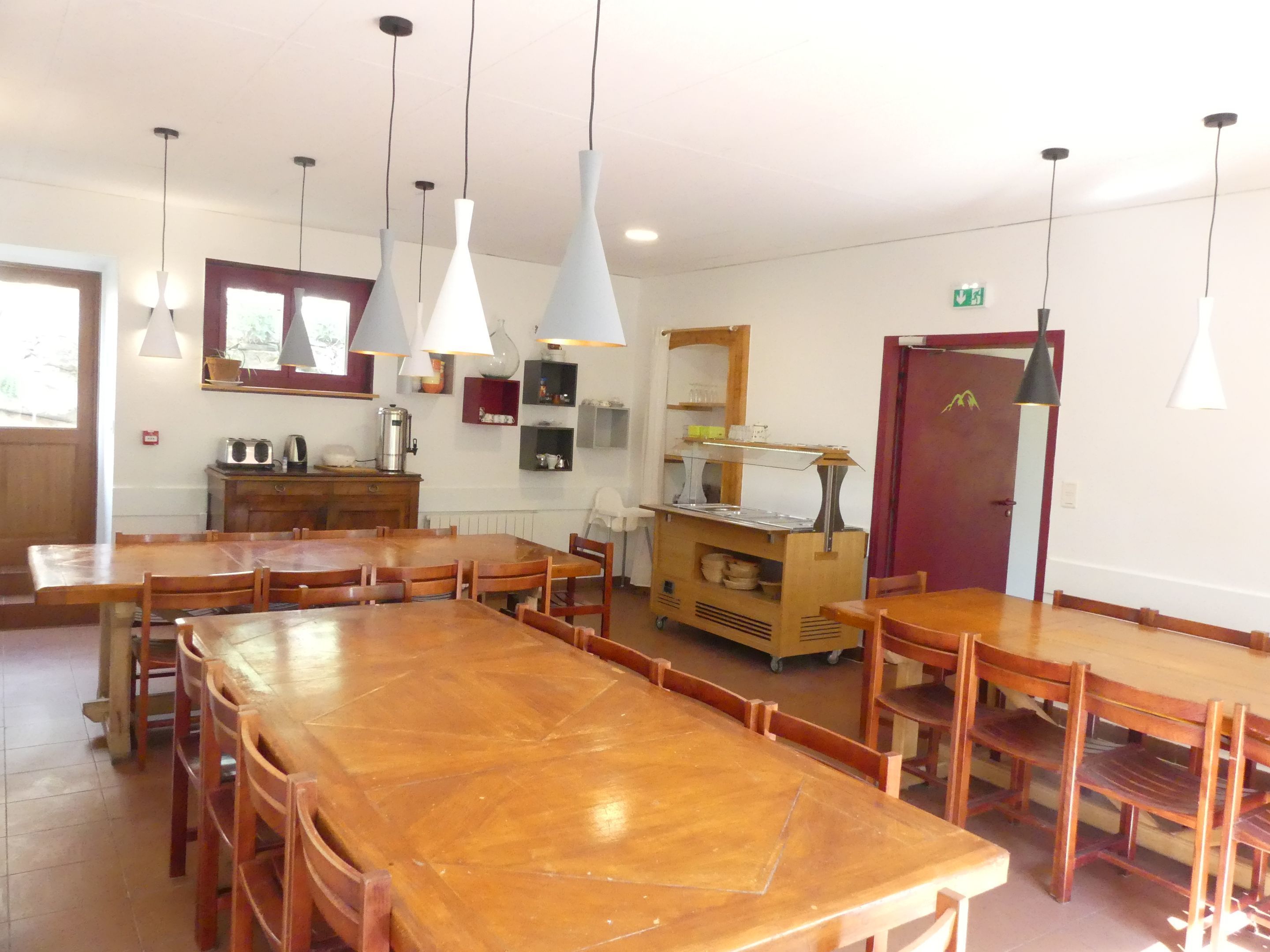 A dining area with wooden tables and chairs, pendant lights, a buffet cart, and a kitchen area with a stove and shelves.