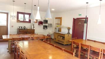 A dining area with wooden tables and chairs, pendant lights, a buffet cart, and a kitchen area with a stove and shelves.