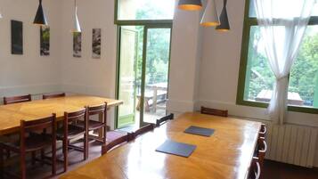 A dining area with wooden tables and chairs, pendant lights, and a view of the outdoors through large windows.