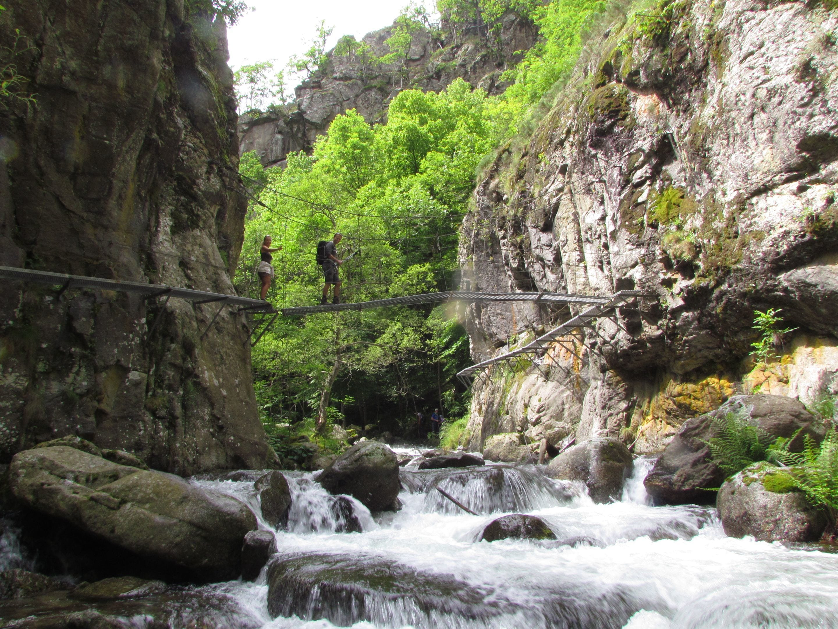 Un puente colgante sobre un río de fuerte corriente, con acantilados rocosos y abundante vegetación.