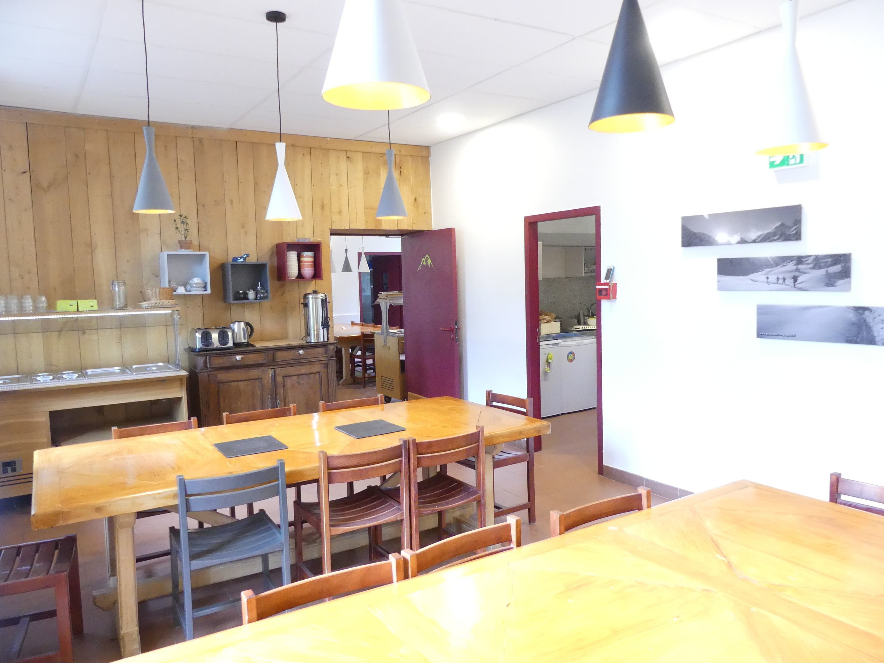 A dining area with wooden tables and chairs, pendant lights, a kitchen area with appliances, and a refrigerator.