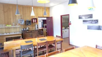 A dining area with wooden tables and chairs, pendant lights, a kitchen area with appliances, and a refrigerator.