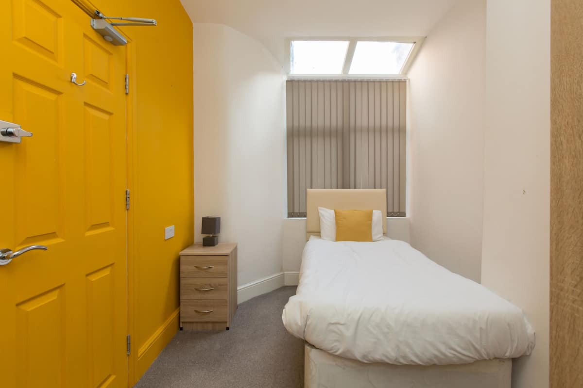 Ein Schlafzimmer mit einem Einzelbett, einem Nachttisch, einer Kommode, einem Fenster mit Vorhängen und einer gelben Tür.