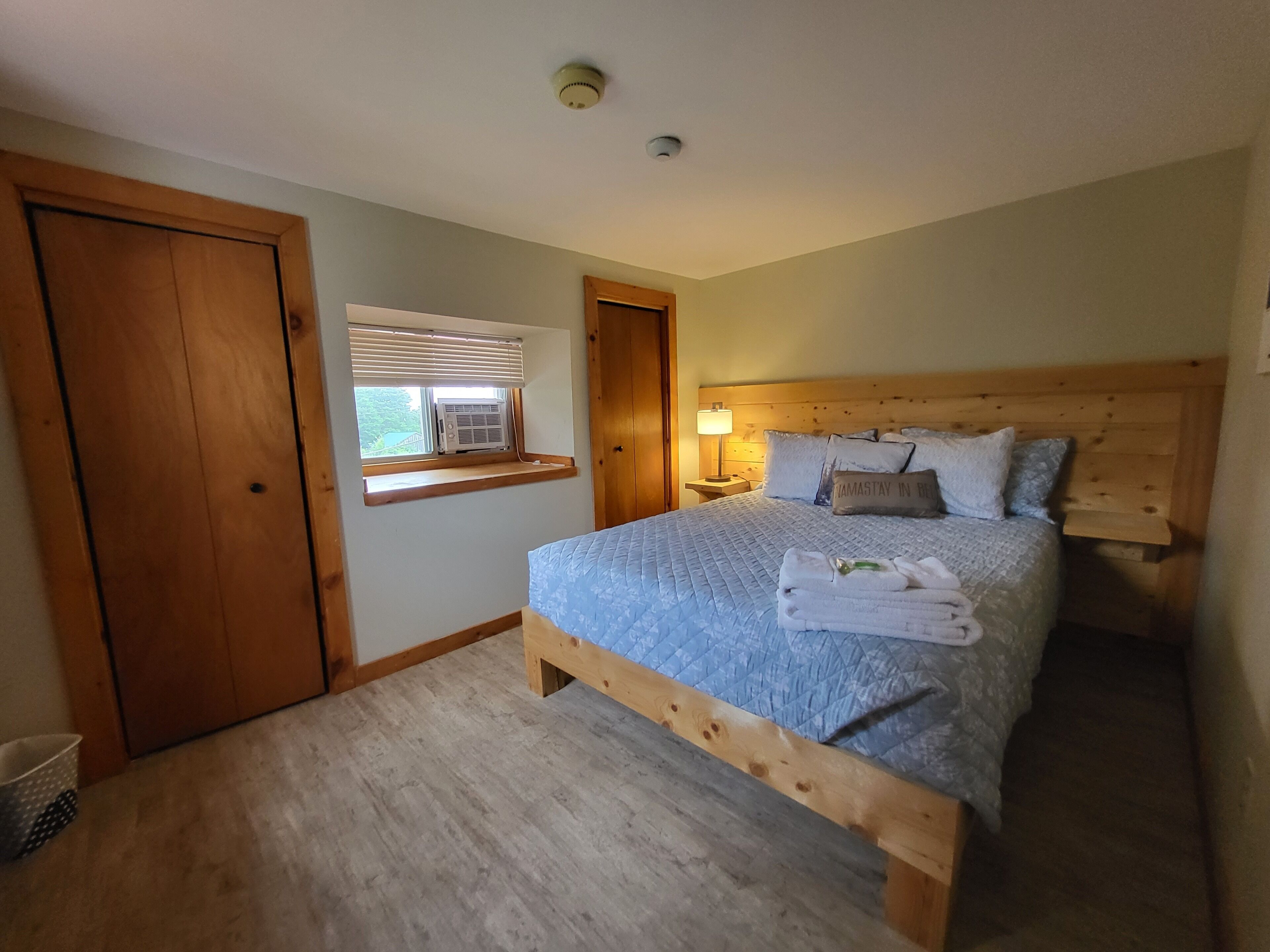A bedroom with a wooden bed frame, a window with blinds, an air conditioning unit, and a nightstand with a lamp.