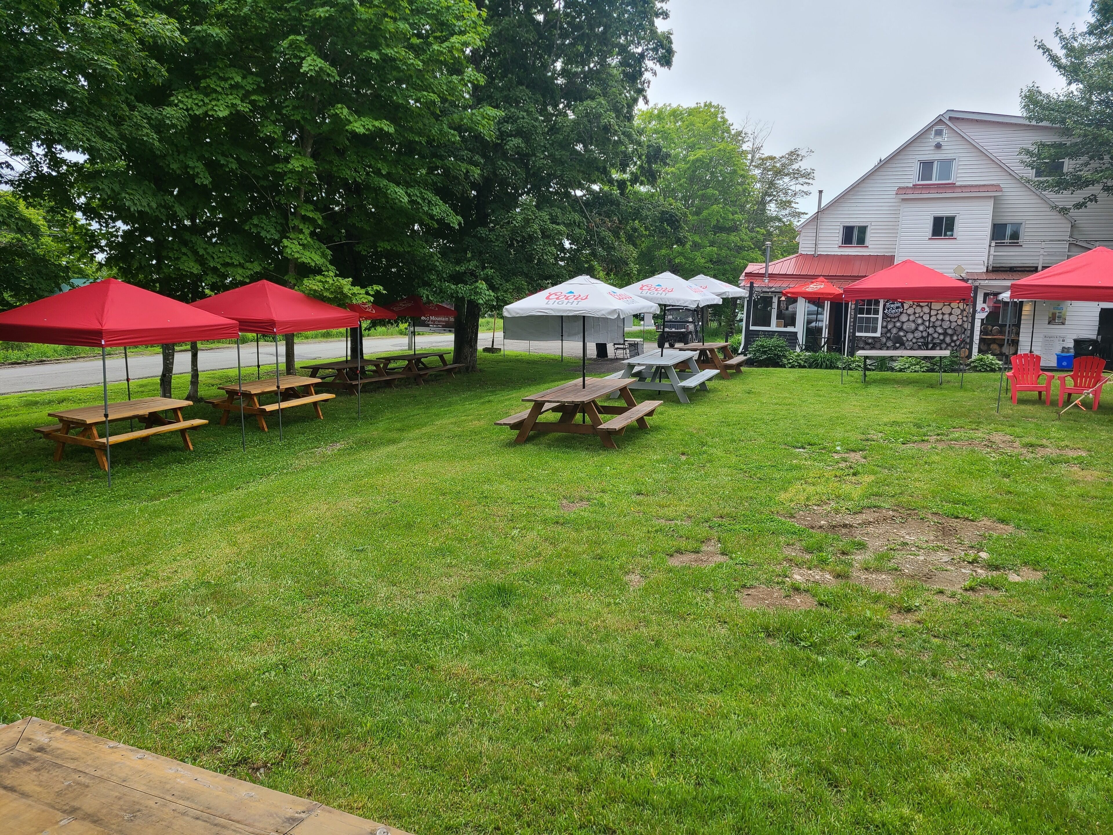 A grassy outdoor area with picnic tables and red umbrellas, a building with a stone facade, and a road in the background.