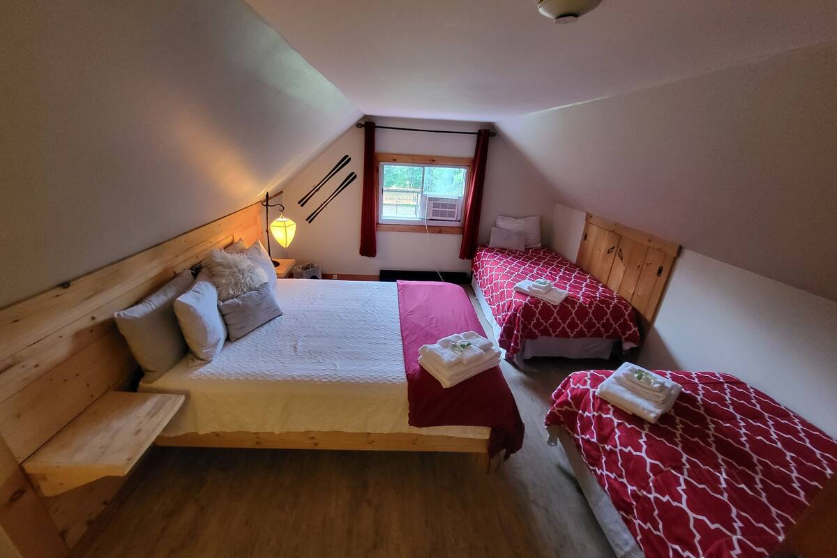 A bedroom with two beds, wooden headboards, a window with curtains, and a small bedside table with a lamp.