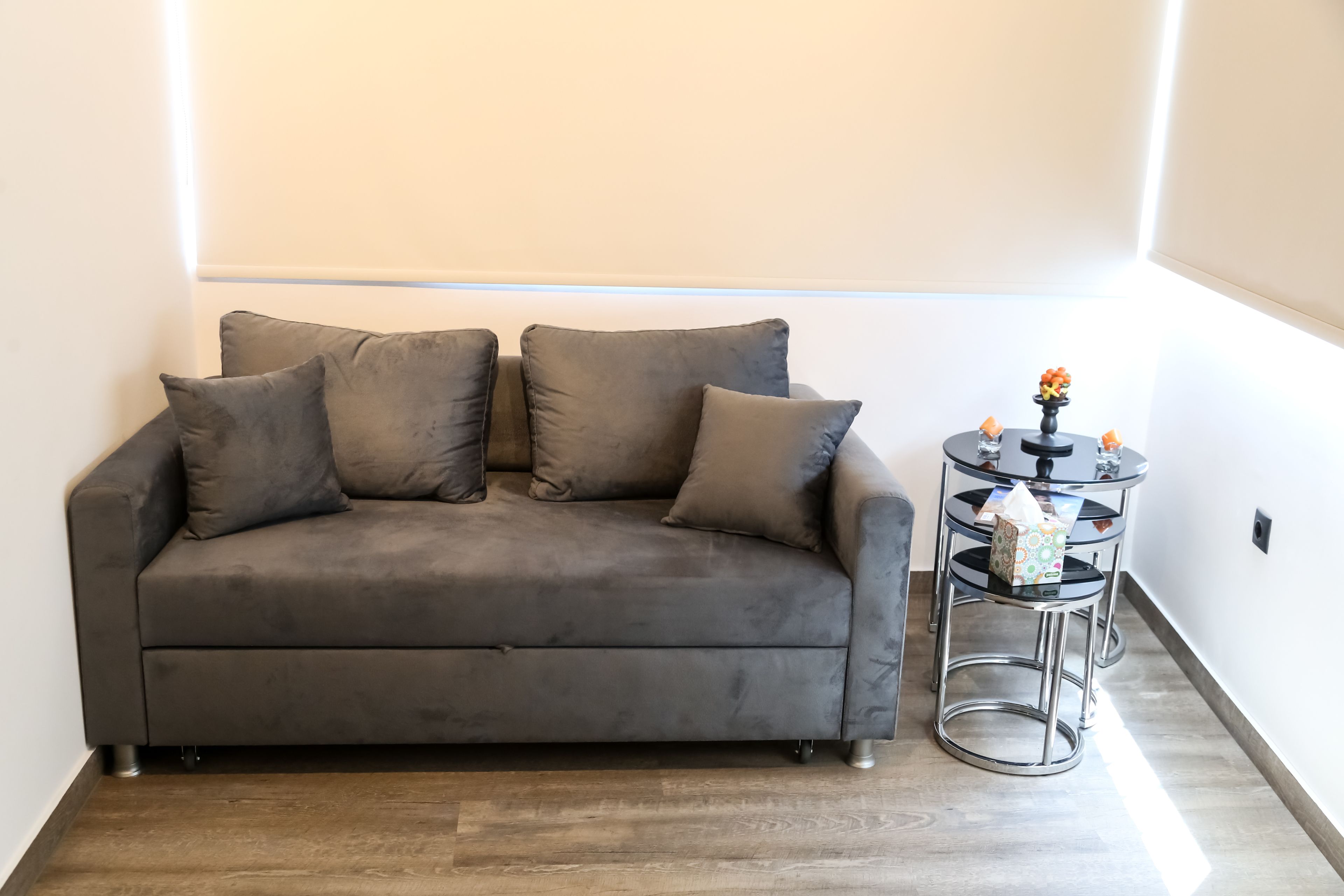 A grey sofa with two cushions, a small round table with a vase and a tissue box, and a wooden floor.